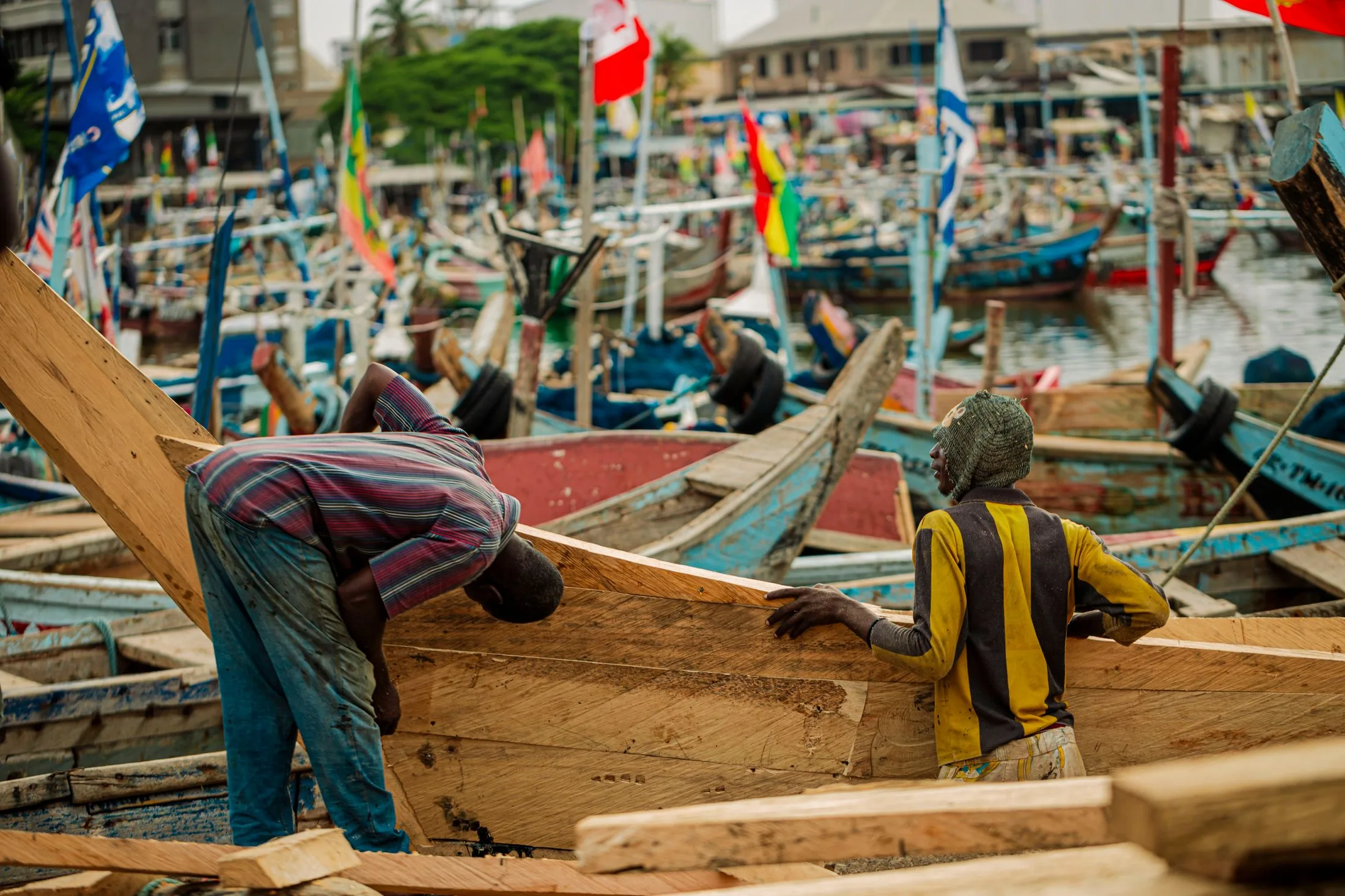 Two boatbuilders fitting a timber plank to a wooden fishing boat at a Ghanaian harbour, with painted boats and Ghanaian flags behind them.