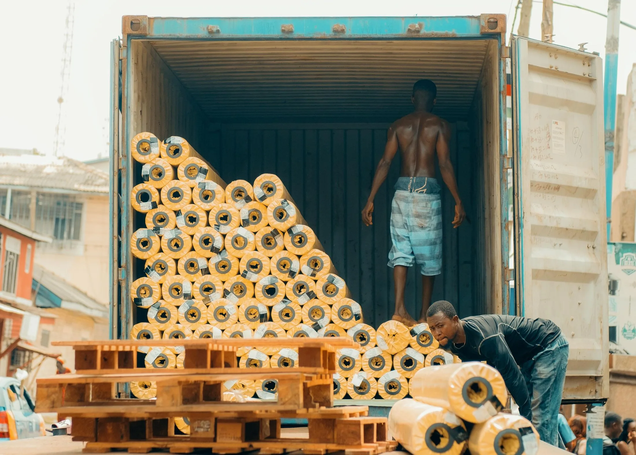 Two workers loading rolls of fabric into a shipping container on a street in Kumasi, Ghana.